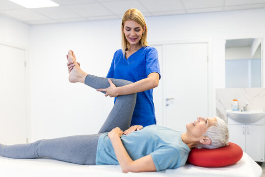 Smiling senior woman performing leg stretching exercises with a female physical therapist in a clinic. Professional rehabilitation and geriatric care for joint mobility and active aging.