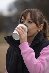 Wrapped in warm clothes, a young woman enjoying a hot drink from a disposable cup finds solace in the cool autumn weather while relaxing in a park surrounded by nature. Vertical photo