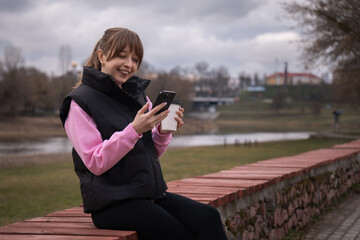 Cheerful young woman is using smartphone and drinking coffee outdoors in city park near river enjoying mobile technology and takeaway beverage on overcast day