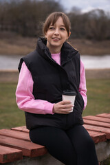 Smiling young woman in pink sweatshirt and black vest sitting on wooden bench holding paper cup with hot drink in her hands and enjoying the fresh air by the river on a cloudy day