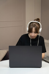 Young woman wearing headphones, working from home while focusing on tasks on her laptop. Surrounded by a modern and comfortable interior, she embodies the essence of remote work productivity