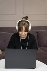 Young woman wearing headphones, working from home while engaged in a video call on her laptop, sitting comfortably on a sofa in her cozy living room, enjoying a relaxed atmosphere