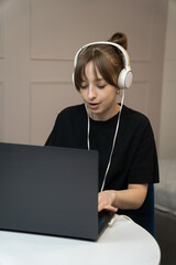 Focused young woman wearing headphones, working diligently on a laptop at a white table in a modern apartment. Vertical photo