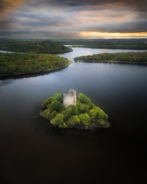 Aerial view of a castle ruin nestled on a small island surrounded by a dark lake, enveloped by a forest under a cloudy sky, Cavan, Ireland.