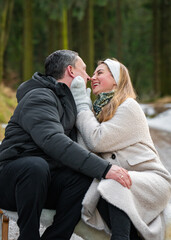 Fototapeta premium Romantic happy man and woman sitting on sled, gently hugging, looking into each other eyes on snowy forest path. Couple in love enjoying winter vacation outdoors, tenderness