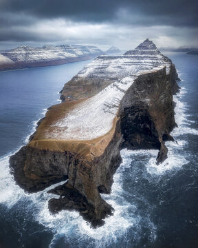 Aerial view of a dramatic peninsula, its snow-dusted peaks contrasting with the dark, rugged cliffs pounded by the frothy, turbulent sea, Trollanes, Northern Isles, Faroe Islands.