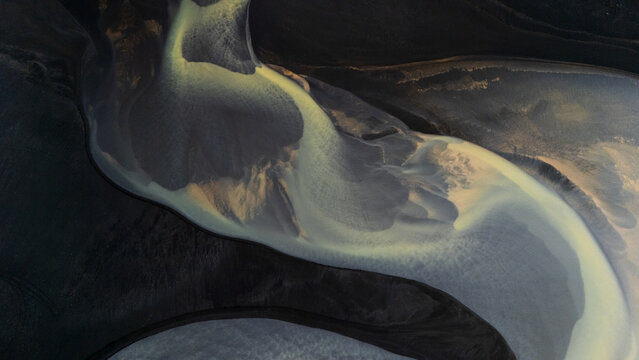 Aerial view of swirling glacial rivers paint an abstract canvas of muted yellows and grays against the stark, dark earth, Selfoss, Iceland.