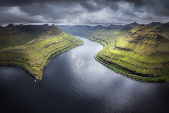 Aerial view of dark waters snake between verdant, striated hills under a brooding sky, Funningur, Eysturoy, Faroe Islands.