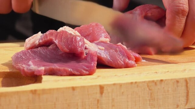 Knife cutting fresh raw pork meat into small pieces on a wooden board. Cooking process in the kitchen. Preparation of ingredients for meal.