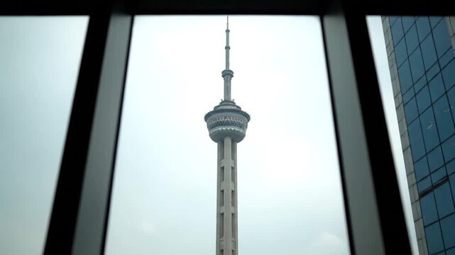 A static, framed view of a prominent transmission tower, likely the Oriental Pearl, against an overcast sky. The tower is centered between dark window frames, flanked by reflections from adjacent mod