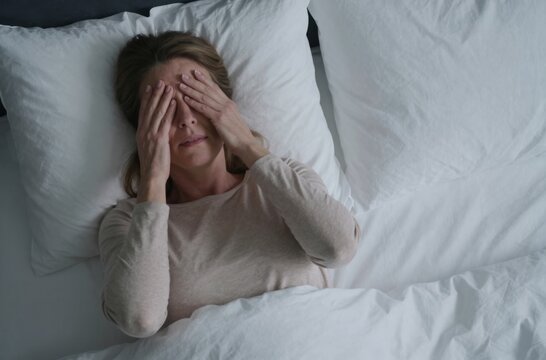Distressed mature woman lying awake in bed at night, covering her eyes due to insomnia, headache, or anxiety and inability to sleep.