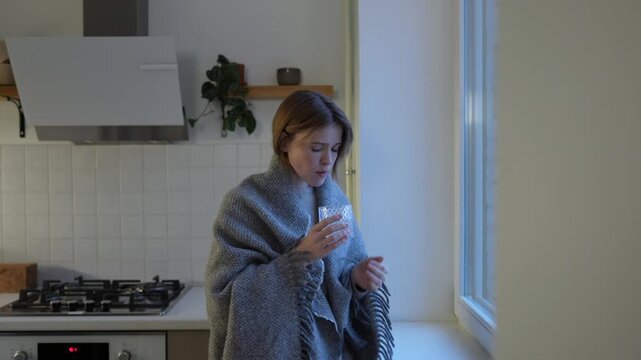 Anxiety sad woman wrapped in wool blanket stands by window, taking pill with water. Emotional exhaustion, psychotherapy. Lost depressed female feeling emotionally drained, swallowing antidepressants.