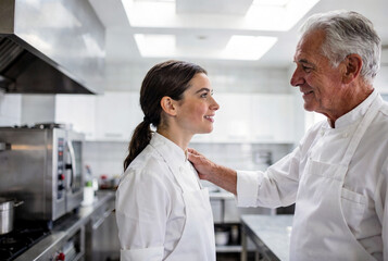 Obraz premium Senior male chef in clean white apron places encouraging hand on shoulder of young female apprentice in bright modern professional kitchen with natural diffused lighting and shallow depth of field. Hi