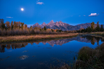 Fototapeta premium A high-resolution landscape of the Teton Range reflected in a calm river during twilight, featuring vibrant autumn foliage and a bright moon in a deep blue sky.