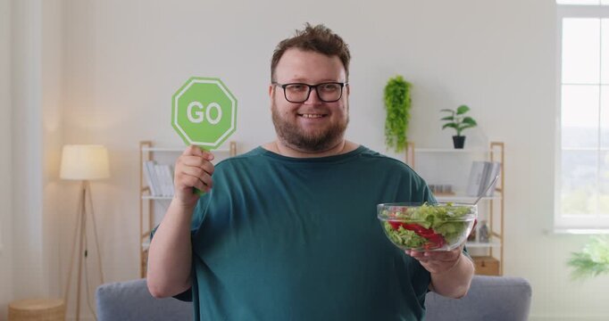 Cheerful fat overweight man at home holds a green sign and bowl of salad, promoting healthy diet, motivation, weightloss, and wellness in living room. Simple message: choose greens and go.
