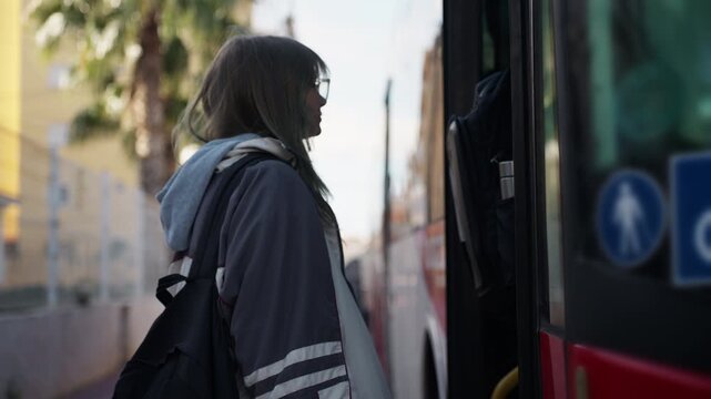 Young female student with glasses and backpack wait at bus stop, tapping card on  contactless reader to pay for ticket, and get on public transport