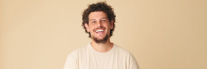 Fototapeta premium Happy guy with curly hair dressed in beige t-shirt laughing looking at camera isolated on beige background in studio. Panoramic
