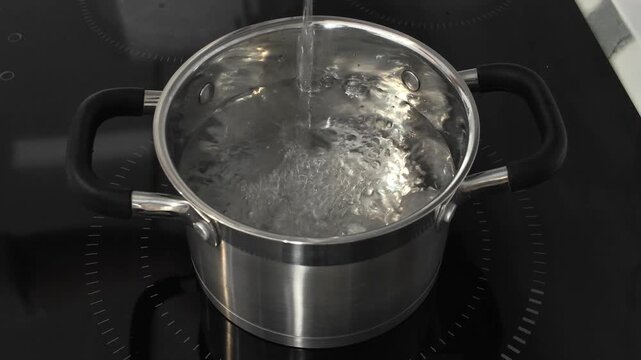 Close-up of clear water pouring into a shiny stainless steel pot. The modern cooking vessel sits on a sleek, black induction stovetop