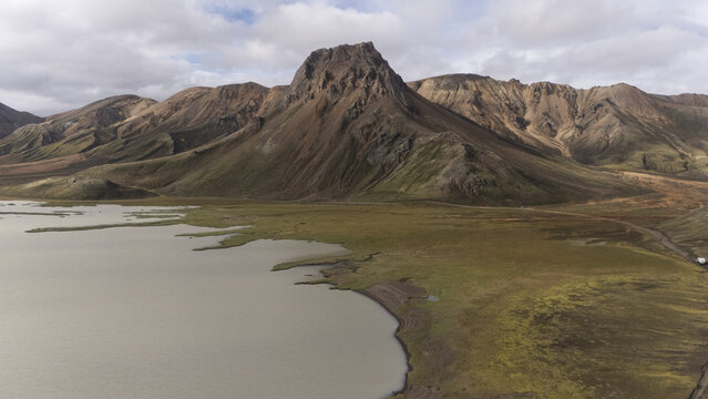 Aerial view of rugged peaks pierce the sky, their ochre slopes cascading into the tranquil lake below, a symphony of earthy tones under a vast, cloudy sky, Selfoss, Iceland.