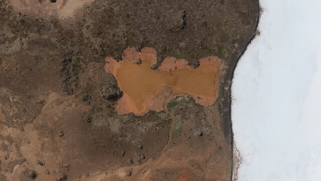 Aerial view of a rusty-red pond nestled amidst the rugged terrain, contrasting sharply with the stark white shoreline, Selfoss, Iceland.