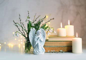 Fototapeta premium praying angel figurine, burning candles, books, snowdrop flowers on table, abstract light background. Christmas or Easter holiday concept. Christian Religious church fest. symbol of faith in God