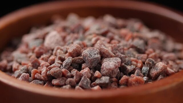 Close-up of black himalayan salt in a brown wooden bowl rotating on a black background.
