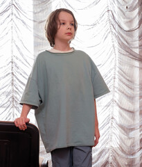 Vertical portrait of a pensive boy in an oversized t-shirt by a window. Child at home