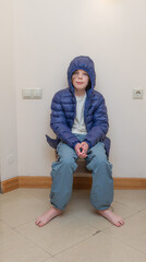 Young boy in a blue hooded down jacket sitting barefoot in a corner. Winter coat indoors