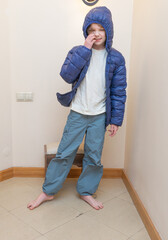 Young boy in a blue puffy jacket with hood up. Standing indoors, smiling and touching his nose. Copy space