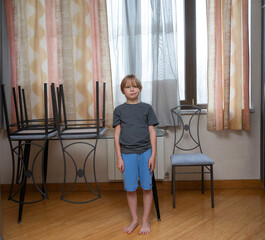 Bored young boy standing barefoot at home. Child with annoyed expression near chairs