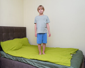 Young boy standing on a bed with green bedding. Portrait of a child in a bedroom