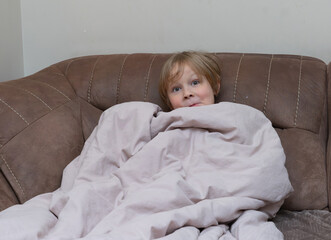 Young boy wrapped in a blanket on a brown sofa. Cute child hiding under a cover at home