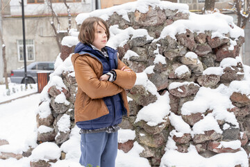 Serious boy with crossed arms in winter snow. Child in warm jacket near rock wall