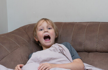 Tired young boy yawning with mouth wide open on a sofa. Sleepy child waking up at home