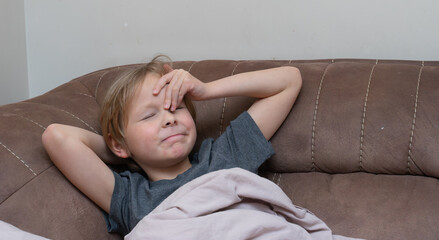 Young boy with a headache touching his forehead on a sofa. Sick child feeling unwell at home