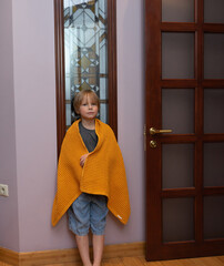 Young boy wrapped in a yellow blanket standing by a door. Child feeling cozy and warm