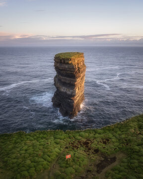 Aerial view of a towering sea stack rising majestically from the turbulent ocean, its layered rock face kissed by the warm glow of the setting sun, County Clare, Ireland.