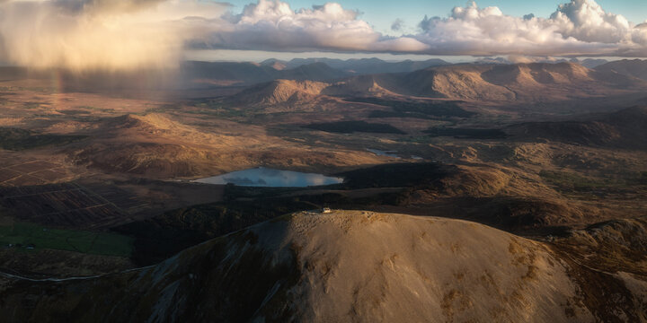 Aerial view of the rugged, sun-kissed peak of Croagh Patrick stands tall against a backdrop of distant mountains and a moody sky, Croagh Patrick, County Mayo, Ireland.
