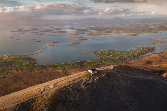 Aerial view of a solitary white building perched atop a rugged peak overlooking the vast expanse of Clew Bay, dotted with islands, Croagh Patrick, Ireland.