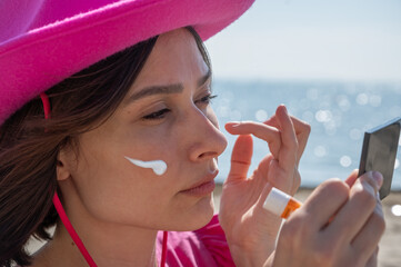 Young woman applying sunscreen and cream for pigmentation spots on her face while walking along the beach. Concept of skincare, sun protection, healthy lifestyle, and outdoor self-care