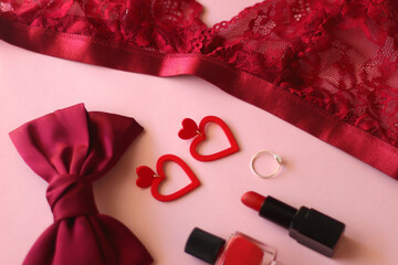 Red lace bralette, hairbow, heart shaped earrings, golden ring, red lipstick and nail polish on pink background. Coquettish accessories for Valentine's day. Selective focus.