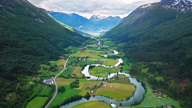 High-angle drone shot following a narrow river through a lush Norwegian valley. Quiet summer mood with scattered farms and dense green hillsides.