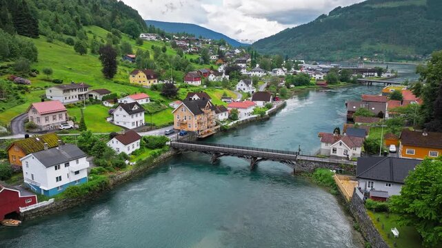 Aerial view of a river bridge in Stryn with colorful houses along the waterfront. Calm summer daylight shows village life framed by green hills and water.