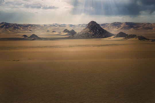 View of sunbeams piercing through the clouds illuminating the rugged, rocky formations rising from the vast, sandy desert landscape, Djanet, Illizi Province, Algeria.