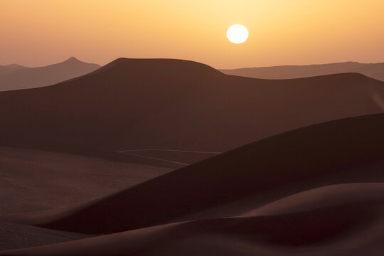 View of the sun blazing over the majestic, undulating sand dunes painted in warm hues of gold and brown, creating a mesmerizing desert landscape, Illizi, Illizi Province, Algeria.