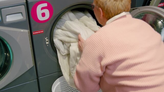 Senior woman in pink sweater loading a large white comforter into a front-loading washing machine at a self-service laundromat with visible numbered machine