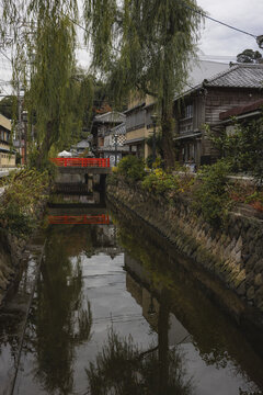 Shimoda, Japan - 25 November 2025: View of a tranquil canal reflecting the vibrant red bridge and the surrounding quaint buildings nestled under weeping willow trees.