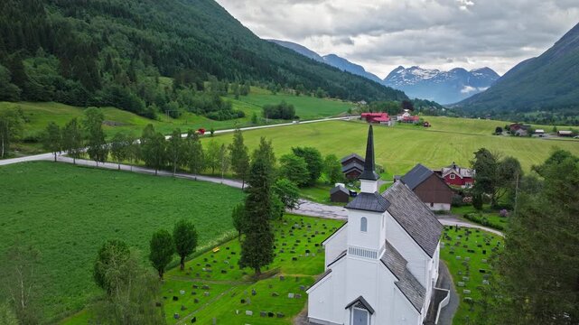 Church and Valley Landscape. Side aerial view of Stryn Church and nearby countryside in Norway, capturing rolling green fields, scattered houses, and mountains in a quiet, pastoral mood.