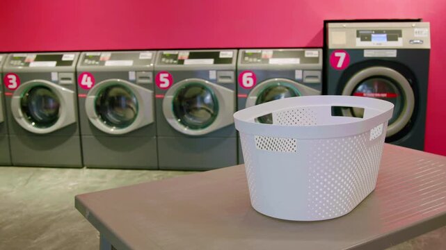 White laundry basket placed on a table in a self-service laundromat with washing machines lined up against a vibrant pink wall, showcasing a modern interior design