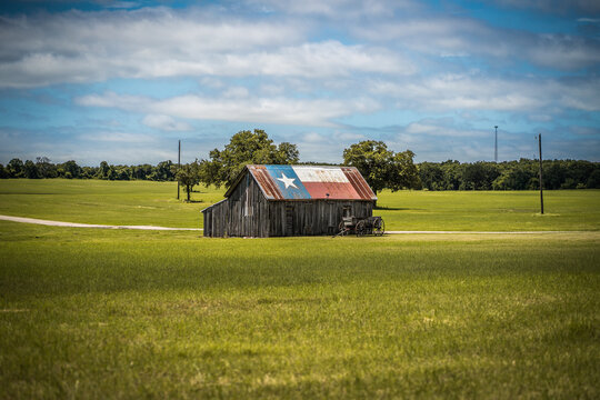 Old Rustic Barn With Texas Flag Painted on Roof With Old Wagon Nearby In Green Field in Rural Town in Texas